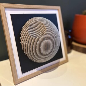 Puede incluir: Una obra de arte 3D enmarcada de una esfera hecha de papel en capas, que se asemeja a un planeta o una luna. La esfera es de color beige claro y se coloca sobre un fondo negro.