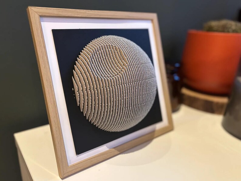 Puede incluir: Una obra de arte 3D enmarcada de una esfera hecha de papel en capas, que se asemeja a un planeta o una luna. La esfera es de color beige claro y se coloca sobre un fondo negro.