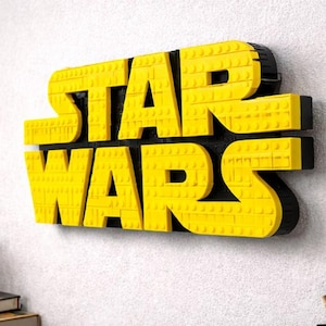 Pode incluir: Letreiro Star Wars amarelo e preto feito de blocos de encaixe. O letreiro est&aacute; montado numa parede branca. Uma pequena planta em vaso e livros s&atilde;o vis&iacute;veis no canto inferior esquerdo.