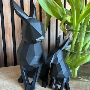 May include: Two black geometric rabbit sculptures, one larger than the other, are sitting on a wooden surface. The sculptures are made of a 3D printed material and have a matte finish.