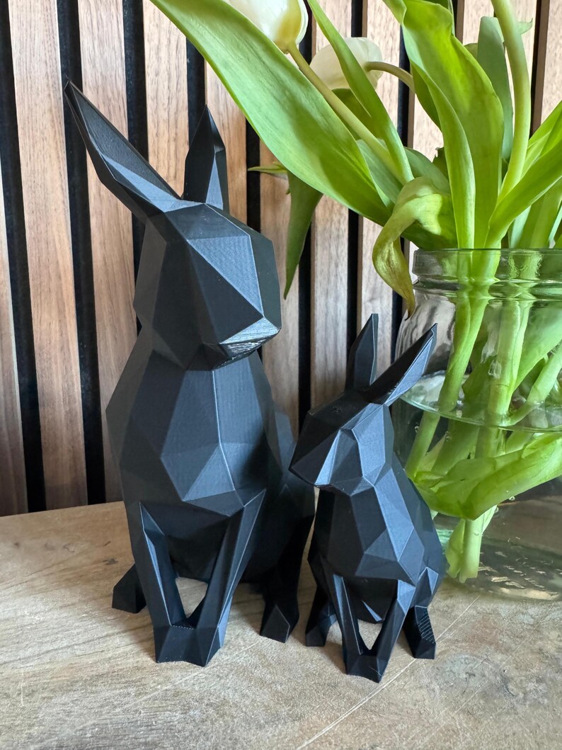 May include: Two black geometric rabbit sculptures, one larger than the other, are sitting on a wooden surface. The sculptures are made of a 3D printed material and have a matte finish.