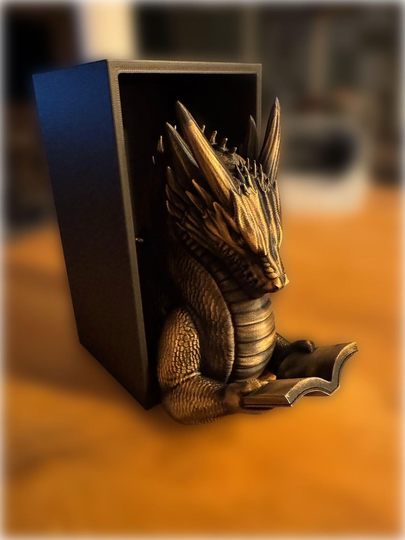 May include: A bronze-colored dragon sculpture reading a book, set within a black rectangular frame. The dragon has detailed scales and horns. The book is held in its front paws. The background is blurred.