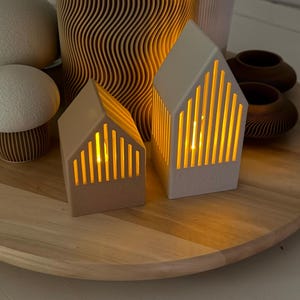 May include: Two small, house-shaped candle holders with vertical cutouts, emitting a warm, yellow glow. They sit on a wooden tray alongside other decorative items, including mushroom-shaped decor and small, dark brown vases.