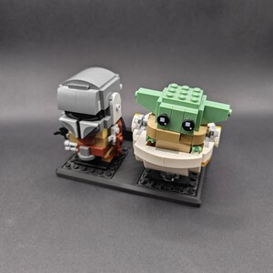 Może przedstawiać: Lego minifigurka Mandalorianina z szarym hełmem i brązową zbroją, stojąca obok Lego minifigurki Grogu, znanego r&oacute;wnież jako Baby Yoda, w białym podzie z zieloną głową i dużymi czarnymi oczami.