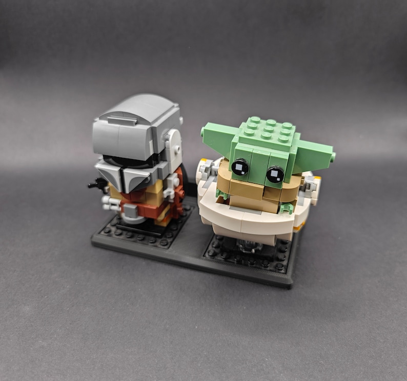 Może przedstawiać: Lego minifigurka Mandalorianina z szarym hełmem i brązową zbroją, stojąca obok Lego minifigurki Grogu, znanego r&oacute;wnież jako Baby Yoda, w białym podzie z zieloną głową i dużymi czarnymi oczami.