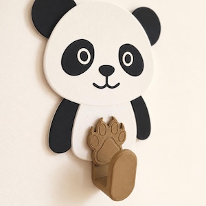 May include: A wall-mounted hook shaped like a panda bear. The panda has a white face, black ears, and black eye patches. A brown, textured paw print design is positioned on the panda's chest, extending into a sturdy, U-shaped hook for hanging.