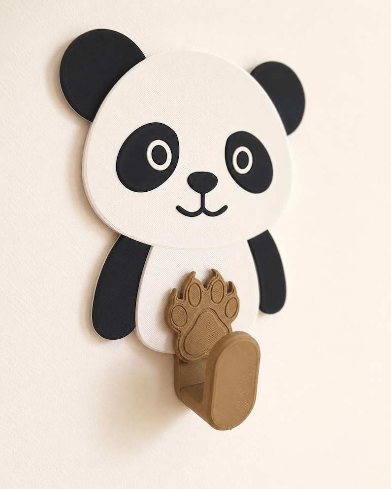 May include: A wall-mounted hook shaped like a panda bear. The panda has a white face, black ears, and black eye patches. A brown, textured paw print design is positioned on the panda's chest, extending into a sturdy, U-shaped hook for hanging.