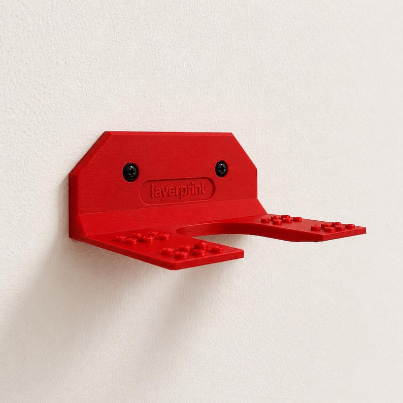 May include: A red plastic wall mount with the text "layerprint" and two black screws. The mount has two arms with raised studs, designed to hold items. The mount is attached to a white wall.