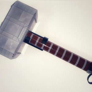 Op de afbeelding: Een grijze en bruine speelgoedhamer, die lijkt op Thor's Mjolnir, gemaakt van in elkaar grijpende blokken. De hamerkop is grijs met rechthoekige panelen. De steel is bruin met grijze strepen en een zwarte riem. De tekst "LEGO INFINITY" is zichtbaar.