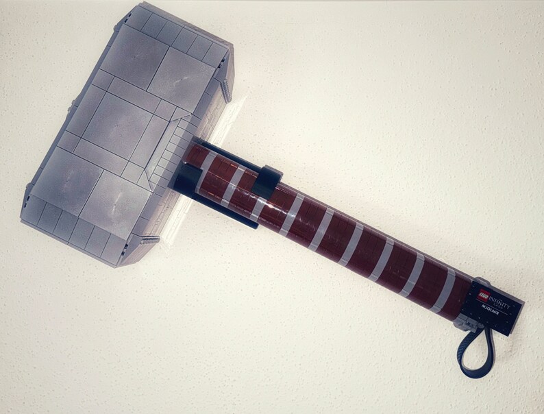 Op de afbeelding: Een grijze en bruine speelgoedhamer, die lijkt op Thor's Mjolnir, gemaakt van in elkaar grijpende blokken. De hamerkop is grijs met rechthoekige panelen. De steel is bruin met grijze strepen en een zwarte riem. De tekst "LEGO INFINITY" is zichtbaar.