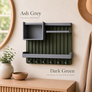 Op de afbeelding: Een wandorganizer met een donkergroen lattenpaneel aan de achterkant en asgrijze onderdelen, waaronder een vierkante opbergdoos, twee horizontale planken en zeven kleine haakjes aan de onderkant. Tekst op de muur leest Ash Grey en Dark Green.
