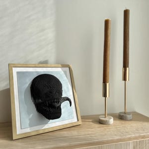 May include: A framed black and white 3D wall art piece depicting a stylized skull with a tongue sticking out. The art is displayed on a wooden shelf with two gold candle holders and brown candles.