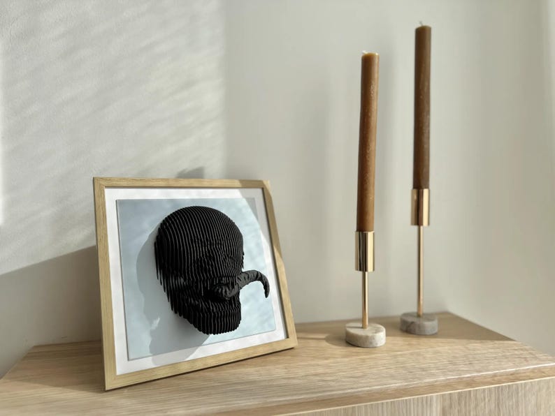 May include: A framed black and white 3D wall art piece depicting a stylized skull with a tongue sticking out. The art is displayed on a wooden shelf with two gold candle holders and brown candles.