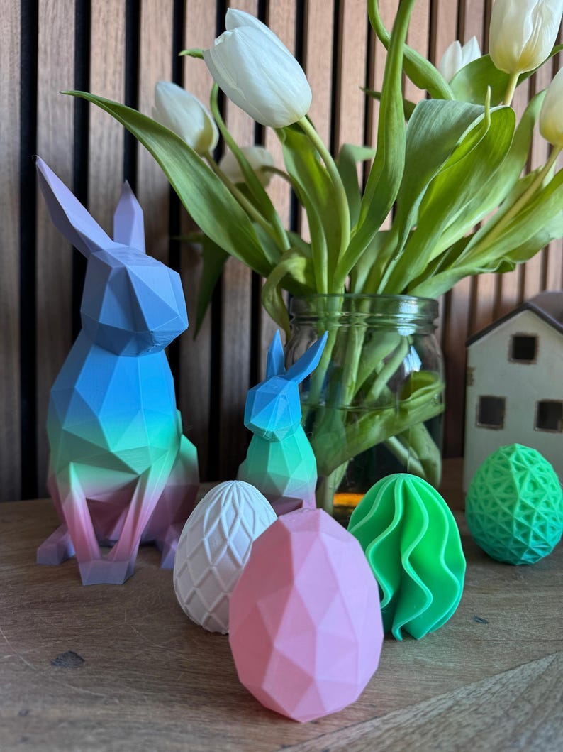 May include: A set of colorful geometric Easter decorations, including a blue and green bunny, a pink egg, a white egg with a textured surface, a green egg with a wavy surface, and a green and white egg with a geometric pattern.