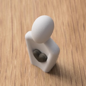 May include: A white abstract sculpture with a textured egg-shaped head and a dark gray heart-shaped element in the chest. The sculpture is set against a wooden background.