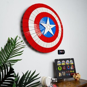May include: A large, circular shield made of red, white, and blue building blocks, resembling Captain America's shield, is mounted on a white wall. A white star is in the center. A display case with collectible pins and a pair of headphones are on a wooden surface.