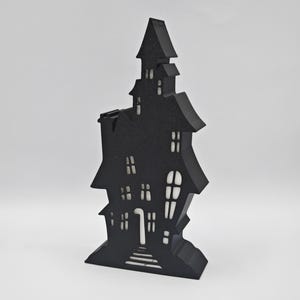 May include: Black silhouette of a haunted house with a white interior. The house has a pointed roof, multiple windows, and a door with steps leading up to it. The house is a Halloween decoration.