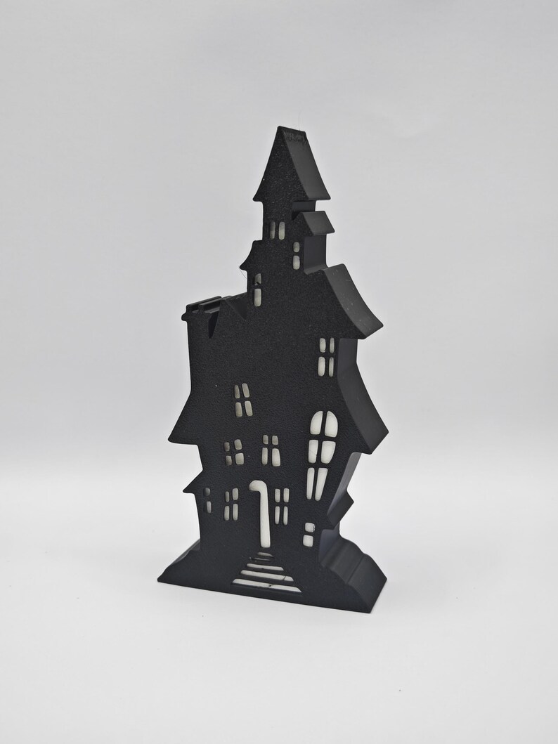 May include: Black silhouette of a haunted house with a white interior. The house has a pointed roof, multiple windows, and a door with steps leading up to it. The house is a Halloween decoration.