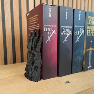 K&ouml;nnte beinhalten: Ein schwarzer, klauenartiger Buchst&uuml;tze h&auml;lt eine Reihe von B&uuml;chern. Die B&uuml;cher tragen Titel wie "Elfen Light" und "Die Elfen" von Bernhard Hennen. Die B&uuml;cher sind in Rot-, Schwarz- und Blaut&ouml;nen gehalten.