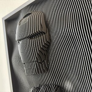 May include: A black and white 3D wall art sculpture of a face, made of layered strips of material. The sculpture is framed and appears to be made of wood or plastic.
