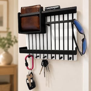 May include: A wall-mounted organizer featuring a white slatted back panel with black trim. It includes a top shelf holding a brown leather wallet and a small black box, a middle shelf for sunglasses, and a bottom row of hooks holding keys and a carabiner.
