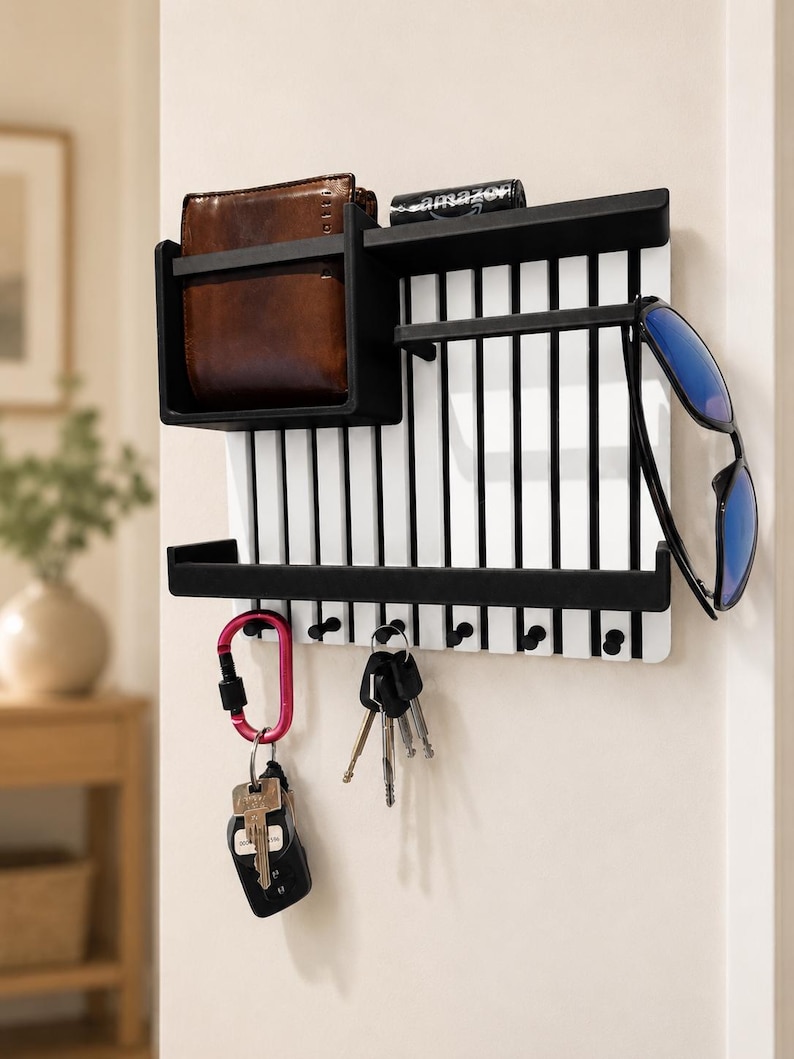 May include: A wall-mounted organizer featuring a white slatted back panel with black trim. It includes a top shelf holding a brown leather wallet and a small black box, a middle shelf for sunglasses, and a bottom row of hooks holding keys and a carabiner.