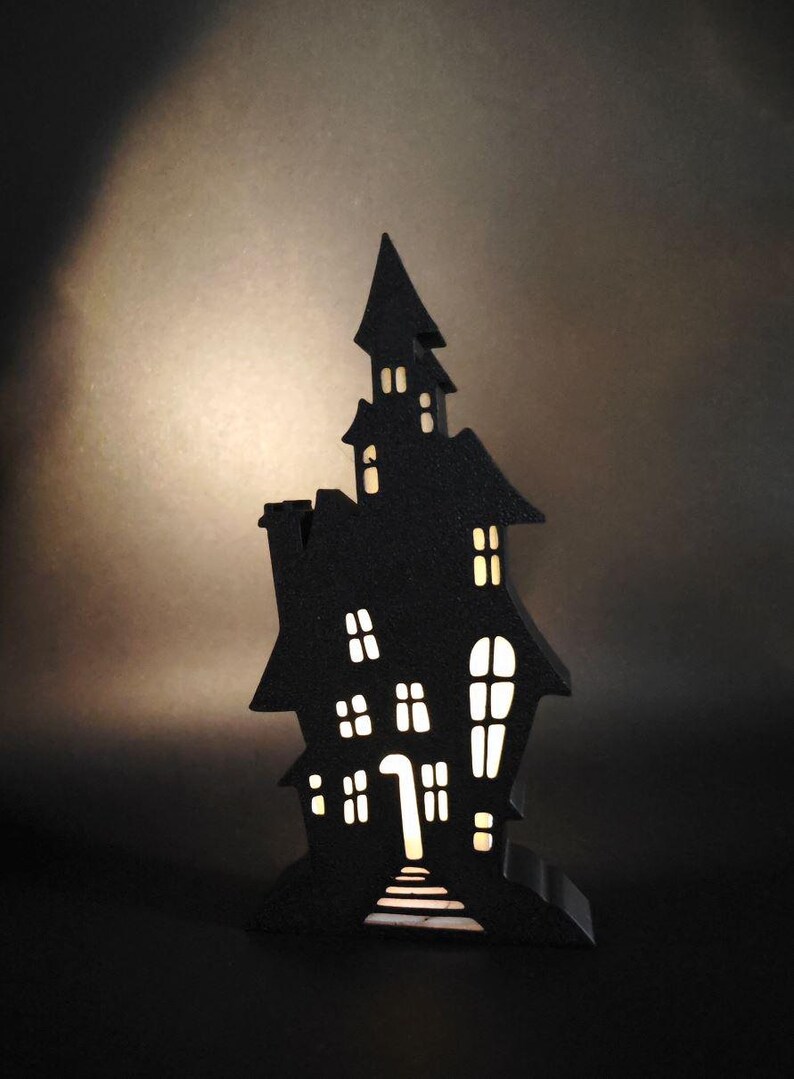 May include: A black wooden haunted house silhouette with cut-out windows and a lit interior. The house is standing on a black surface.