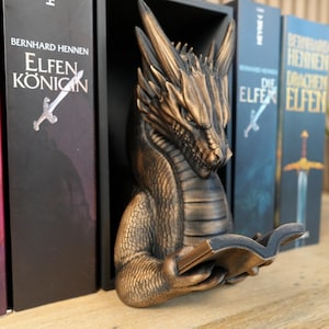 May include: A bronze-colored dragon bookend, holding a book, sits between several fantasy novels. The dragon has detailed scales and a focused expression. The book titles are in German.