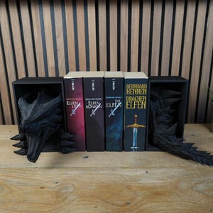 May include: A black dragon-themed bookshelf with four books. The books have titles in German. The bookshelf has a dragon head on the left and a dragon tail on the right. A small potted plant sits to the right of the books.
