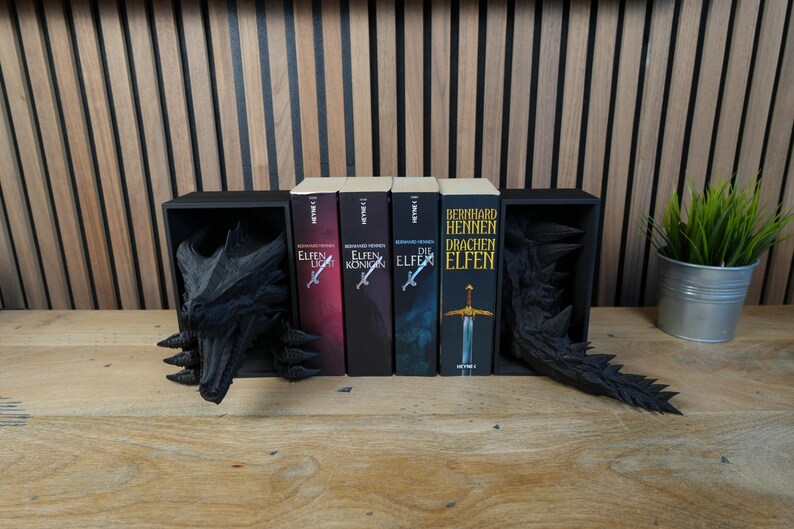 May include: A black dragon-themed bookshelf with four books. The books have titles in German. The bookshelf has a dragon head on the left and a dragon tail on the right. A small potted plant sits to the right of the books.