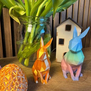 May include: Two geometric origami-style bunny rabbits, one orange and one with a gradient of blue, green, and pink. The bunnies are sitting on a wooden surface with a vase of green tulips in the background.
