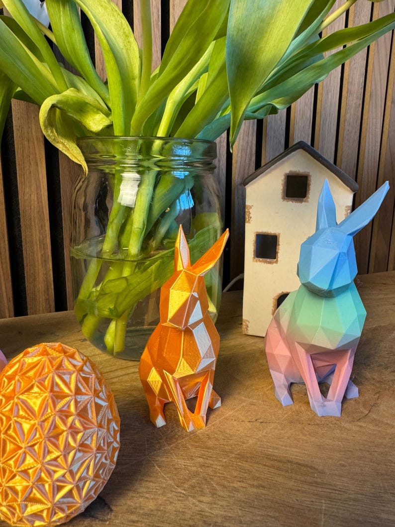 May include: Two geometric origami-style bunny rabbits, one orange and one with a gradient of blue, green, and pink. The bunnies are sitting on a wooden surface with a vase of green tulips in the background.