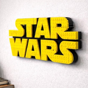 Pode incluir: Letreiro amarelo e preto "STAR WARS" feito de blocos de brinquedo interligados. O letreiro est&aacute; montado em uma parede branca. Uma pequena planta em vaso e livros s&atilde;o vis&iacute;veis no canto inferior esquerdo.