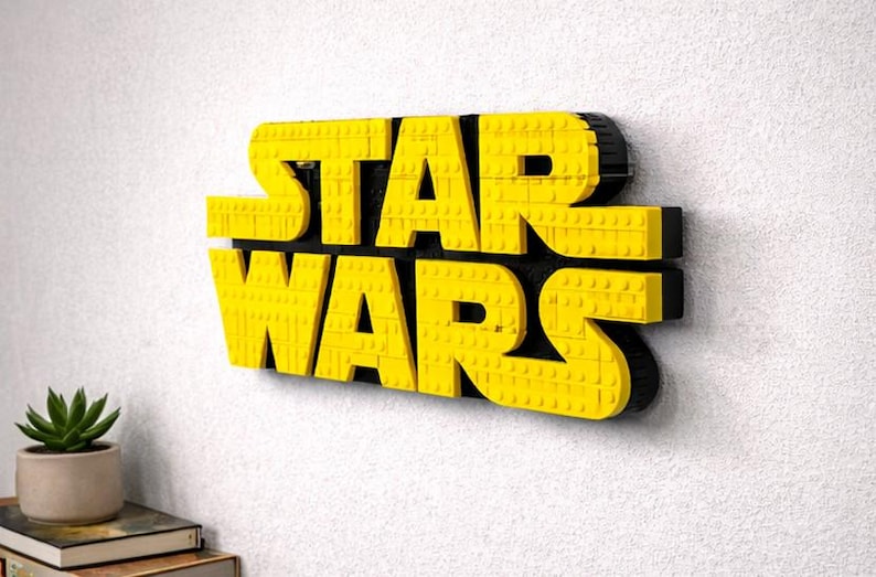 Pode incluir: Letreiro amarelo e preto "STAR WARS" feito de blocos de brinquedo interligados. O letreiro est&aacute; montado em uma parede branca. Uma pequena planta em vaso e livros s&atilde;o vis&iacute;veis no canto inferior esquerdo.