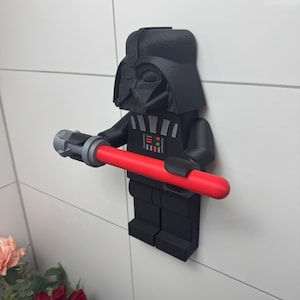 May include: A black and grey 3D printed Darth Vader LEGO minifigure holding a red lightsaber. The minifigure is mounted on a white tiled wall.