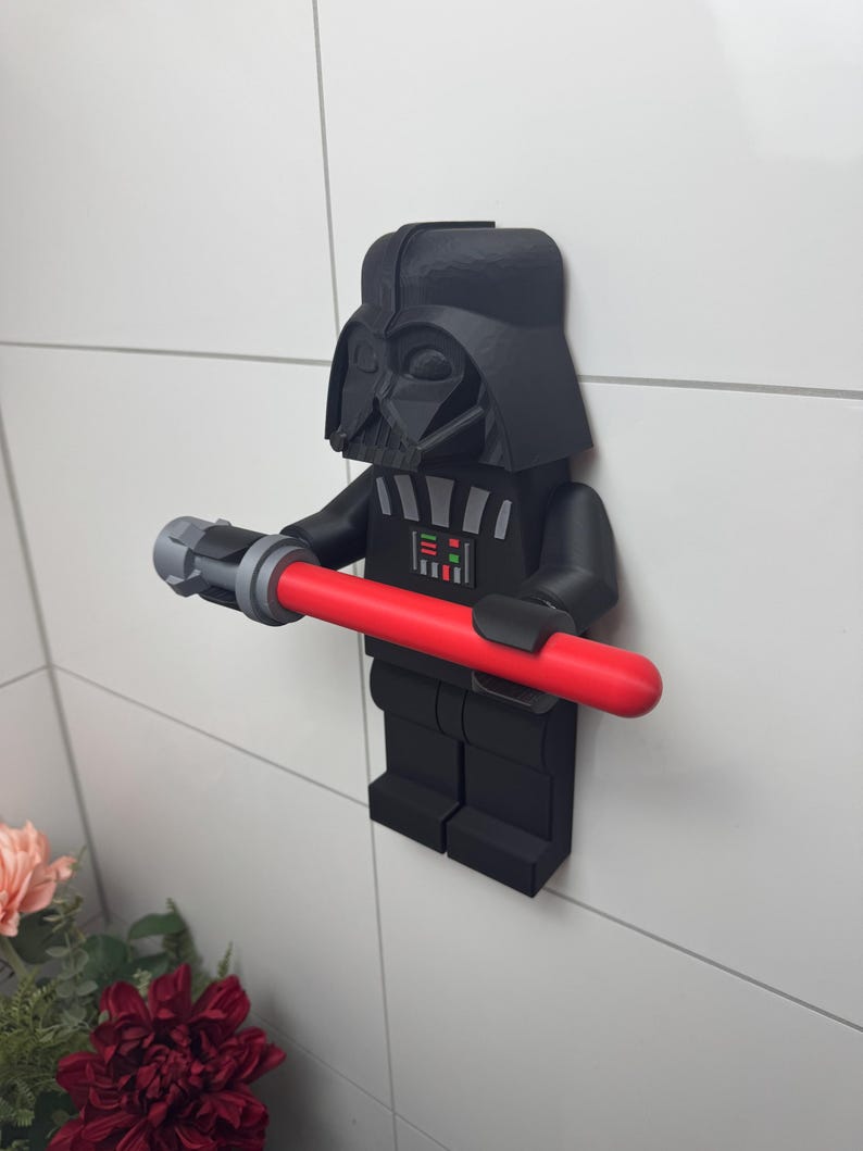 May include: A black and grey 3D printed Darth Vader LEGO minifigure holding a red lightsaber. The minifigure is mounted on a white tiled wall.