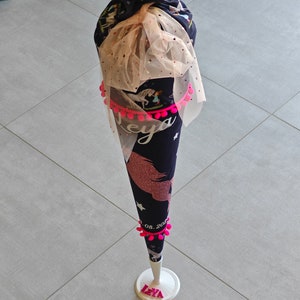 Puede incluir: Un cono escolar de unicornio azul y rosa con pompones rosas y la fecha "08.2023" impresa en &eacute;l. El cono est&aacute; en un soporte blanco con el nombre "Leya" impreso en letras rosas.