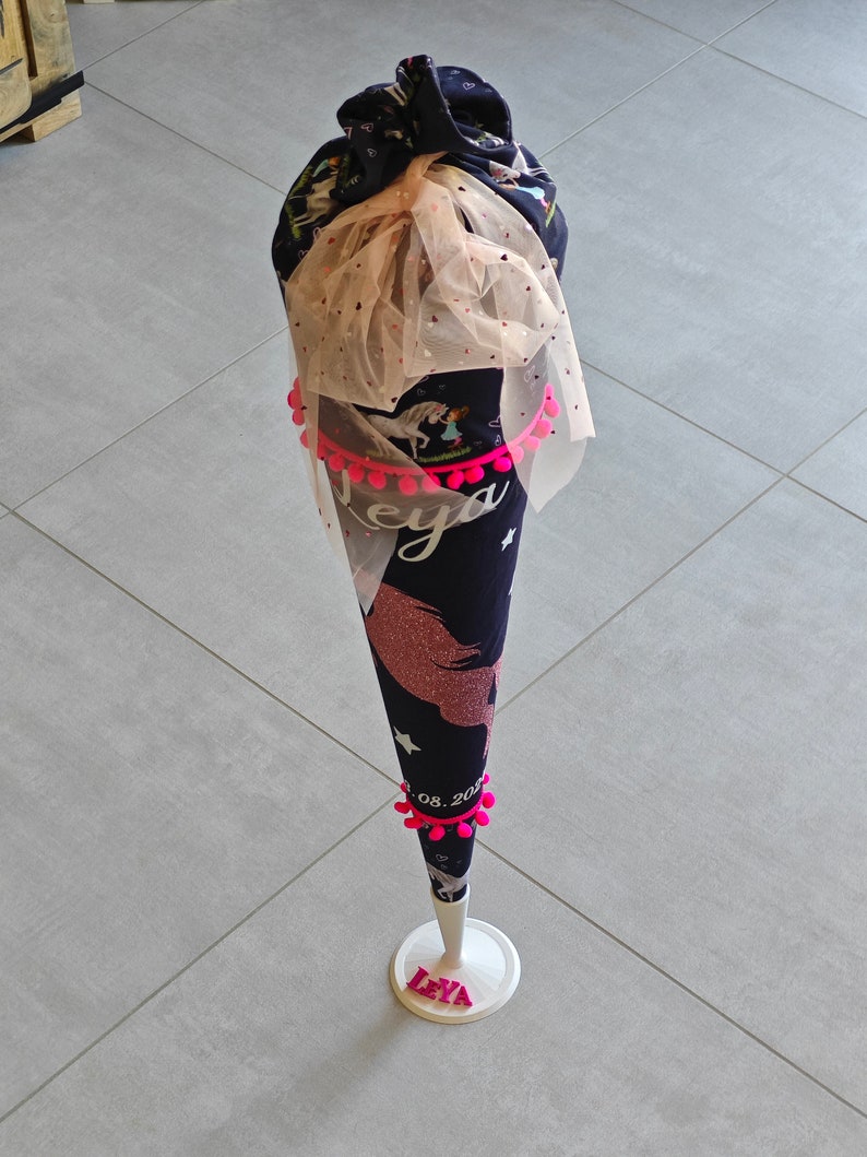 Puede incluir: Un cono escolar de unicornio azul y rosa con pompones rosas y la fecha "08.2023" impresa en &eacute;l. El cono est&aacute; en un soporte blanco con el nombre "Leya" impreso en letras rosas.