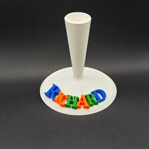 Puede incluir: Soporte blanco impreso en 3D con un soporte en forma de cono y el nombre "RICHARD" escrito en letras azules, verdes, naranjas y rojas.