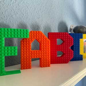 May include: Colorful block letters spelling "FABIO" in green, orange, red, blue, and yellow. The letters are made to resemble building blocks and are displayed on a white shelf against a light blue wall. A decorative astronaut figure is in the background.