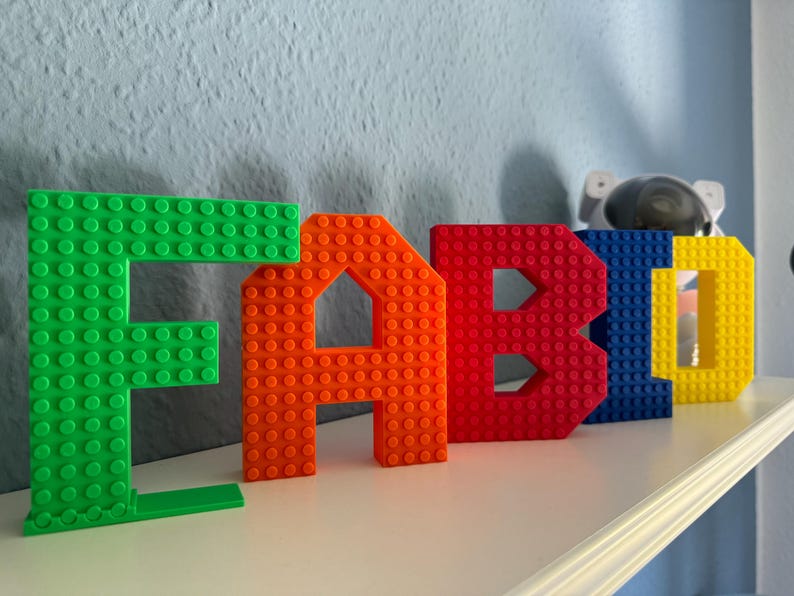 May include: Colorful block letters spelling "FABIO" in green, orange, red, blue, and yellow. The letters are made to resemble building blocks and are displayed on a white shelf against a light blue wall. A decorative astronaut figure is in the background.