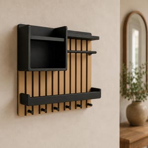 May include: A wall-mounted organizer featuring a light wood slatted back panel with a black storage box, a horizontal bar, a shelf, and seven black hooks along the bottom edge. The unit combines natural wood tones with matte black metal components.