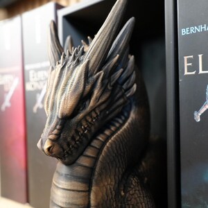 May include: A bronze-colored dragon sculpture with detailed scales and horns, positioned between book spines. The dragon's head is in profile, with a focused expression. The books have titles in German.