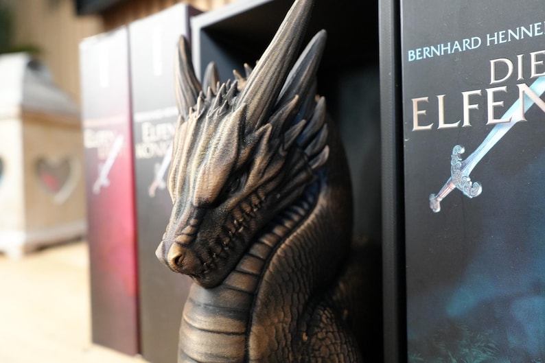 May include: A bronze-colored dragon sculpture with detailed scales and horns, positioned between book spines. The dragon's head is in profile, with a focused expression. The books have titles in German.