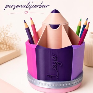 May include: A pencil-shaped desk organizer with a pink, purple, and dark purple exterior featuring the name Leya in script. The top has multiple compartments holding colored pencils, and the base is accented with a silver band and pink bottom.