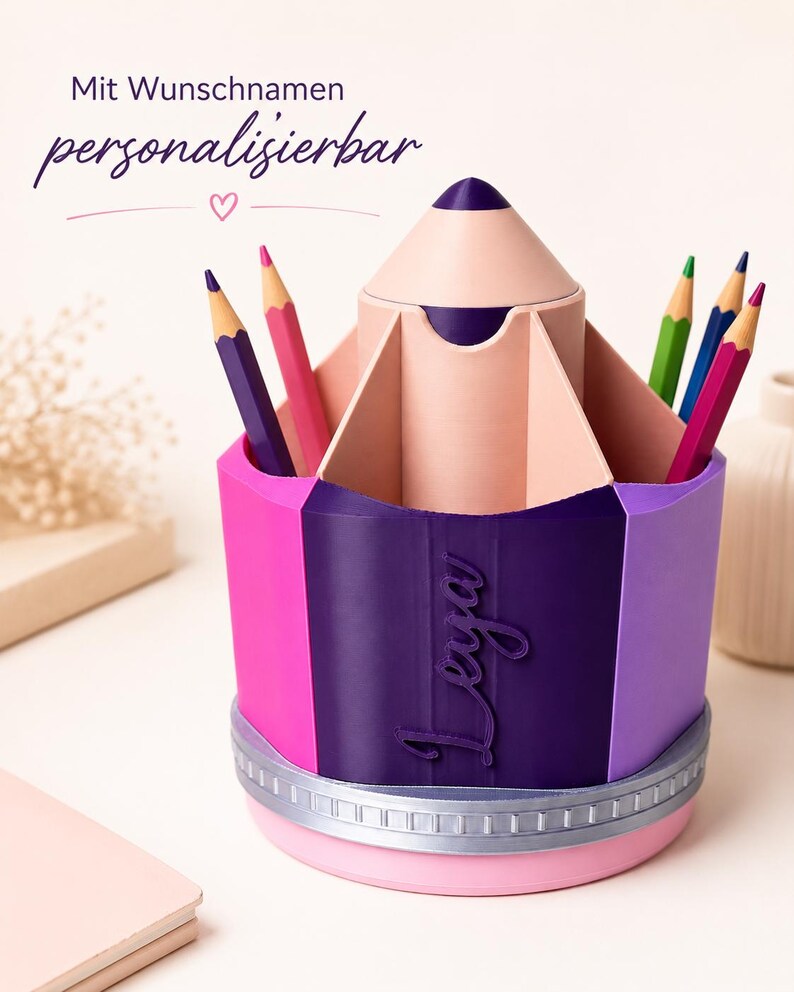 May include: A pencil-shaped desk organizer with a pink, purple, and dark purple exterior featuring the name Leya in script. The top has multiple compartments holding colored pencils, and the base is accented with a silver band and pink bottom.