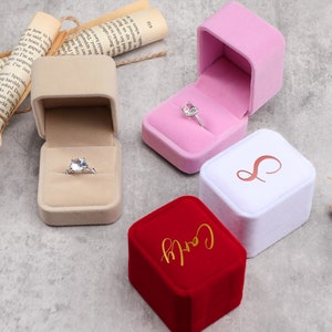 Könnte beinhalten: Drei Ringboxen, eine beige, eine rosa und eine rote. Die beige Box enthält einen silbernen Ring mit einem großen, klaren Stein. Die rosa Box enthält einen silbernen Ring mit einem großen, klaren Stein. Die rote Box hat den Schriftzug "Carly" in goldenen Lettern.