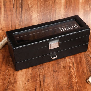 Könnte beinhalten: Schwarze Lederuhrenbox mit Glasdeckel und silbernem Griff. Die Box hat zwei Schubladen und der Name "Driscoll" ist auf der Oberseite eingraviert.