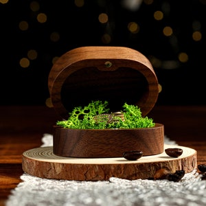 Könnte beinhalten: Eine hölzerne Ringbox mit grüner Moos-Auskleidung und einem goldenen Ring im Inneren. Die Box steht auf einer Holzscheibe mit Kaffebohnen, die um sie herum verstreut sind.