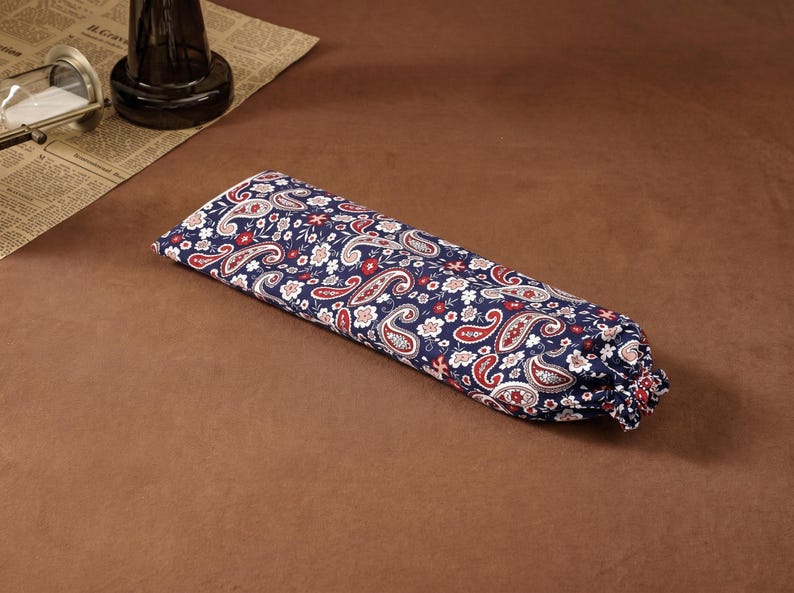 Puede incluir: Una bolsa de tela rectangular con fondo azul oscuro y un patr&oacute;n repetitivo de remolinos de cachemira blancos y rojos junto a peque&ntilde;os motivos florales. La bolsa tiene un extremo fruncido con cierre de cord&oacute;n, creando una forma cil&iacute;ndrica suave.